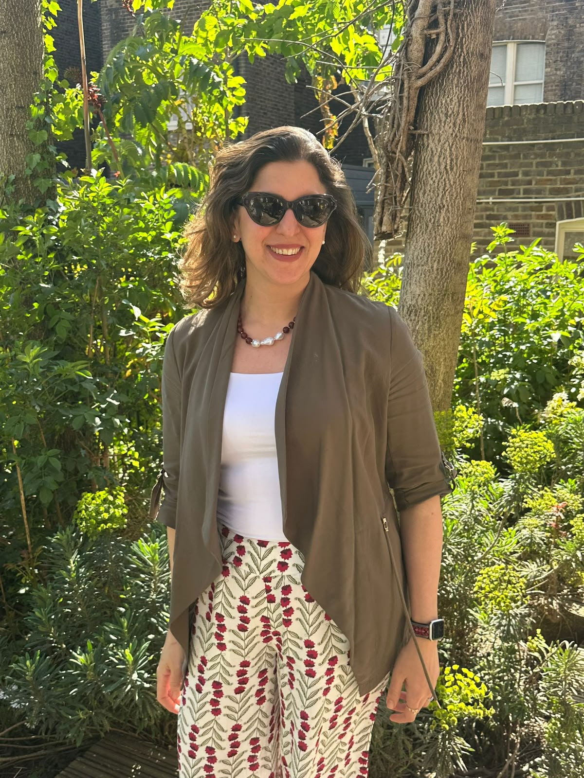 Farah in an olive jacket and floral trousers standing in a garden
